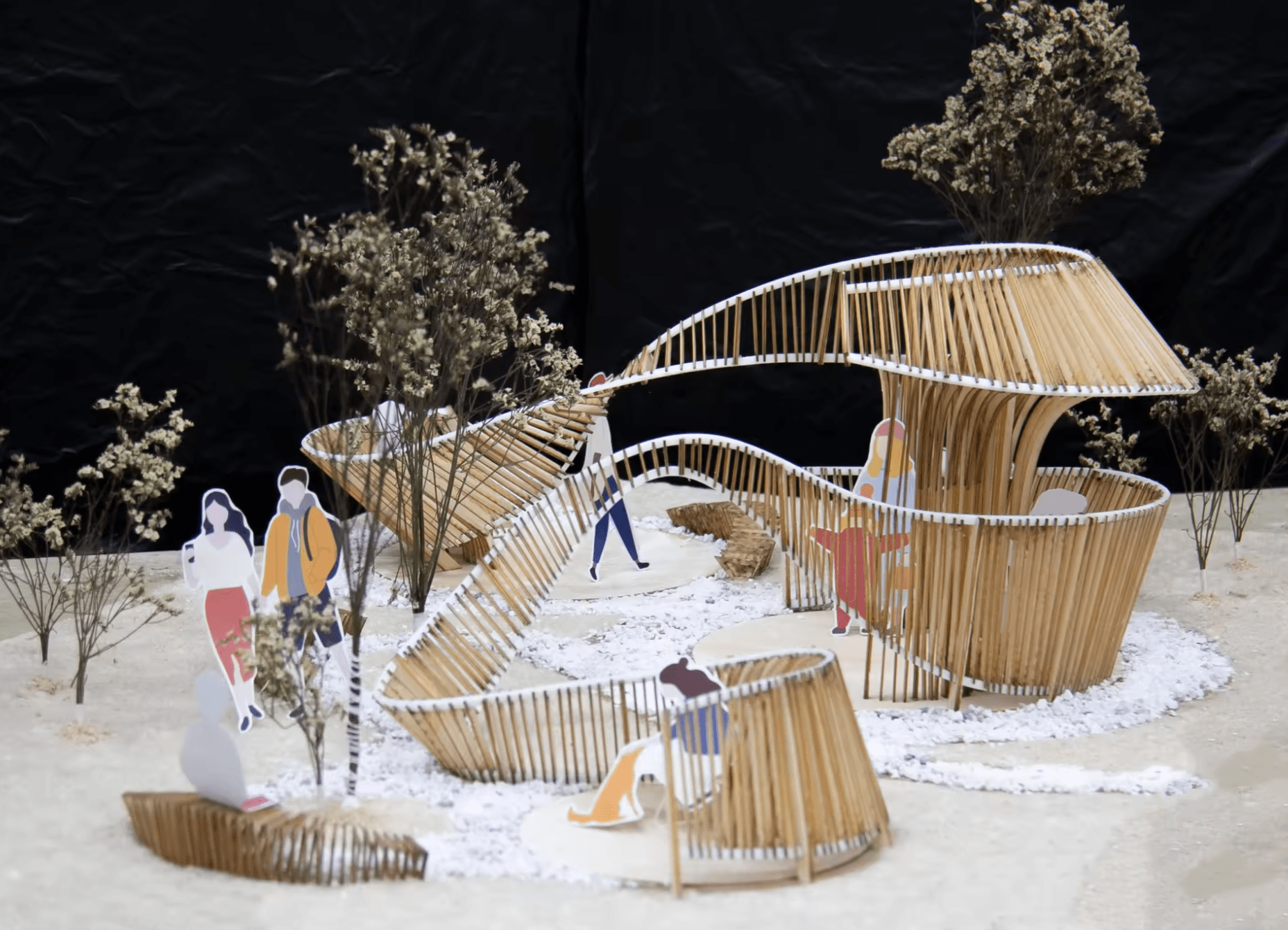 Physical model of a winding ribbon formed by slender vertical pieces, rising and dipping around trees and seated and standing figures